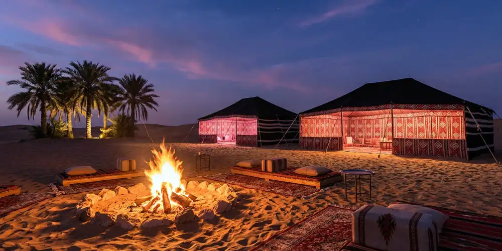 A roaring central campfire surrounded by traditional floor seating and glowing tents under a deep purple twilight sky.