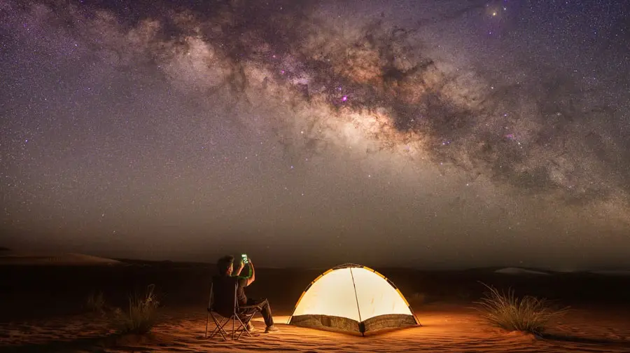 Camper sitting next to an illuminated tent taking photos of the vibrant Milky Way galaxy in the Abu Dhabi desert.