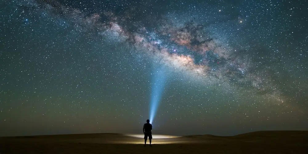 Silhouette of a person standing on desert dunes shining a flashlight beam up into the Milky Way night sky in Abu Dhabi.