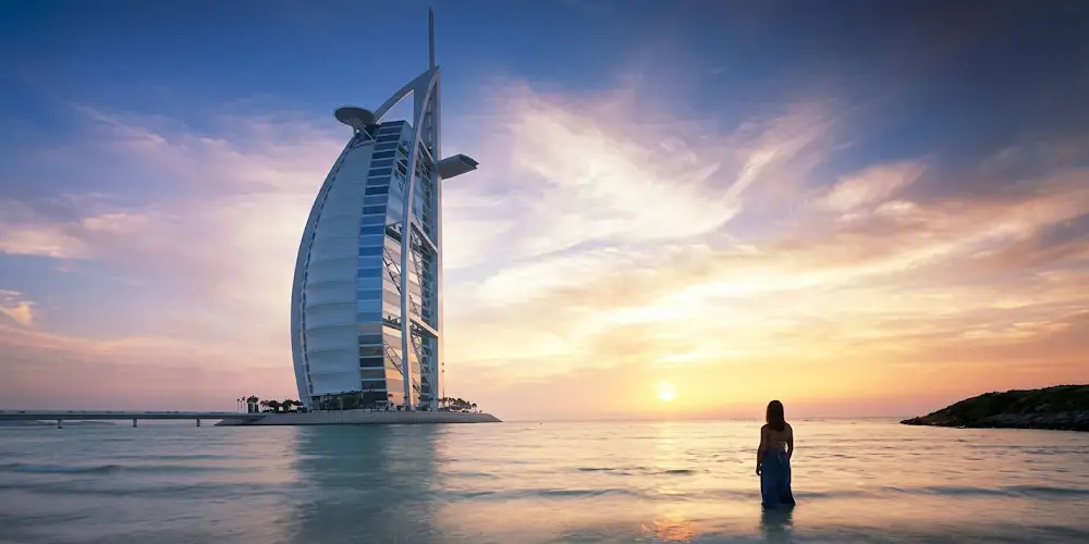 Silhouette of a woman standing in shallow sea water on the beach looking at the iconic Burj Al Arab hotel during sunset in Dubai.
