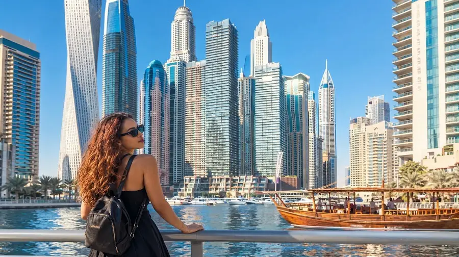 Woman leaning on the railing along Dubai Marina promenade with luxury high-rises and a traditional wooden dhow in the background.