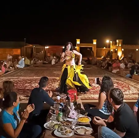 A belly dancer in a yellow and black costume performs on a carpeted stage for a seated audience enjoying dinner at an outdoor desert camp at night.