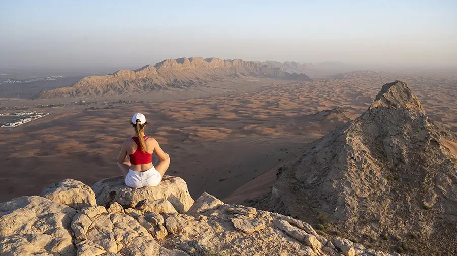 Woman meditating on Fossil Rock in Sharjah overlooking expansive desert plains and mountain ranges.