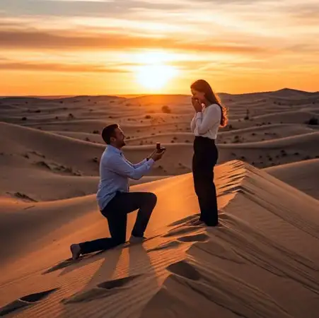 A man kneels to present a ring to a surprised woman on the crest of a sand dune, silhouetted against a vibrant orange sunset.