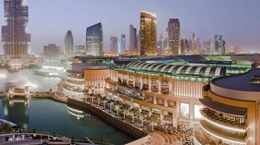 High-angle evening view of The Dubai Mall waterfront promenade and Dubai Fountain lake with surrounding illuminated skyscrapers.