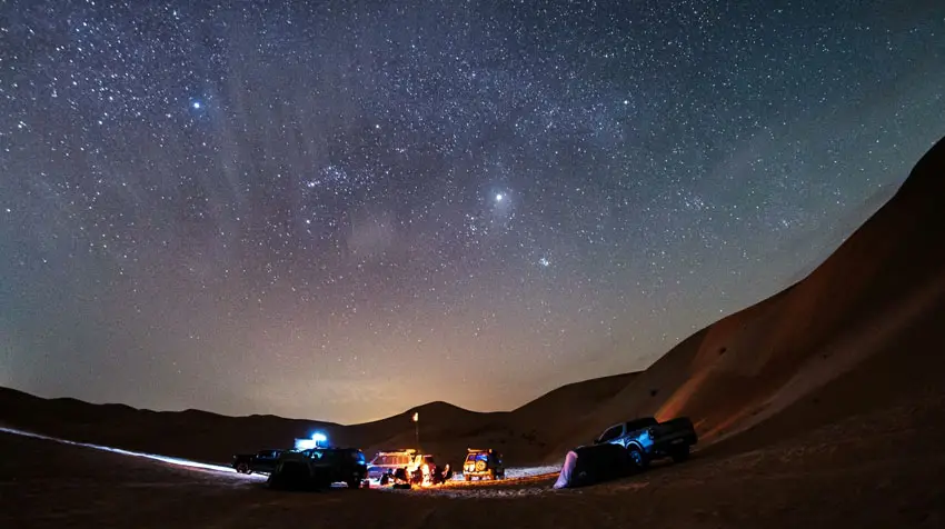 Off-road vehicles and camping tents set up around a campfire under a starry night sky at Al Quaa Desert in Abu Dhabi.