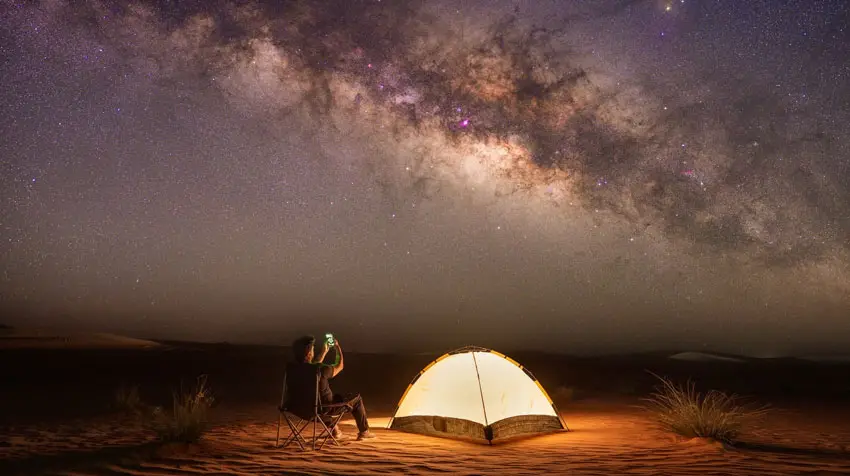 Stargazer sitting by an illuminated tent taking photos of the bright Milky Way core in Al Quaa Desert, Abu Dhabi.