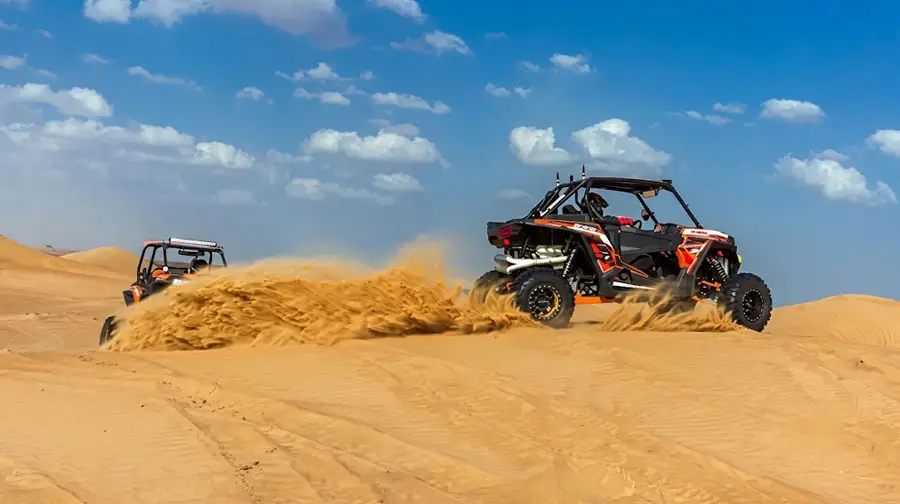 Two orange and black dune buggies speeding over golden sand dunes in Abu Dhabi, kicking up large clouds of sand under a bright blue sky during an exciting desert tour.