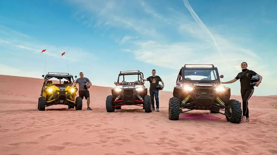 Three powerful dune buggies (yellow, red, and dark) parked in the Abu Dhabi desert with their drivers posing proudly in front, headlights on, ready for an exciting off-road desert safari.