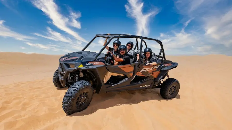 A happy group of four friends riding together in a black and orange Polaris RZR dune buggy in the vast sand dunes of Abu Dhabi, waving and enjoying their desert tour under a blue sky.