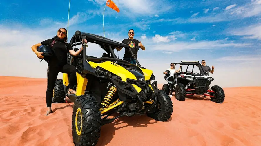 A group of smiling tourists posing with a bright yellow and black dune buggy in the Abu Dhabi desert, one woman holding a helmet while others stand near additional vehicles under a clear blue sky.