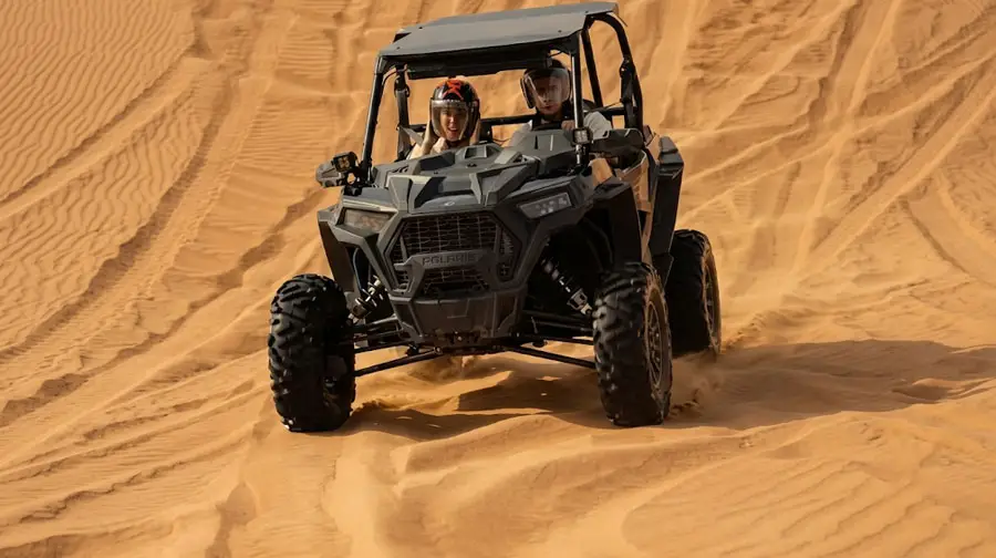 A gray Polaris RZR dune buggy with a male driver and female passenger navigating steep sand dunes in Abu Dhabi, captured mid-action during an exhilarating desert adventure tour.