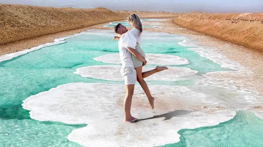 Couple posing playfully on white salt crust formations standing over emerald green water at Al Wathba Salt Lake in Abu Dhabi.