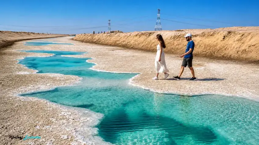 Man and woman strolling along the shoreline of turquoise water channels at Al Wathba Salt Lake in Abu Dhabi.