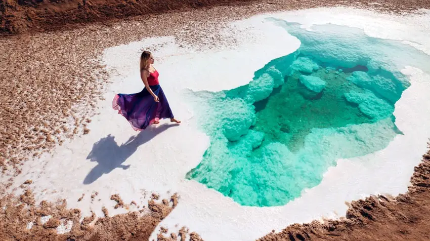 High-angle view of a woman in a flowing dress walking along the edge of a crystal-clear turquoise salt pool at Al Wathba Salt Lake in Abu Dhabi.