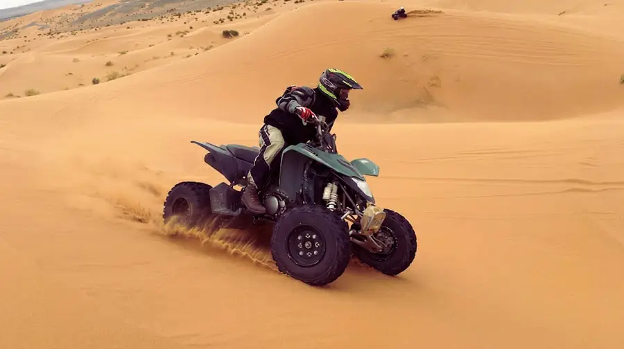 Adventurer in black gear and neon helmet riding a green and black quad bike up a steep sand dune in the Abu Dhabi Desert, kicking up a trail of golden sand with another rider visible in the distance