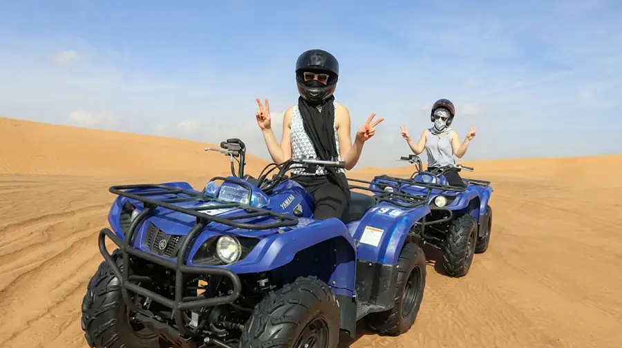 Two excited women on blue Yamaha quad bikes posing with peace signs in the Abu Dhabi Desert sand dunes, both wearing helmets and enjoying their desert adventure.