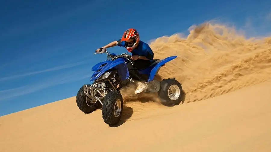 Thrilling action shot of a rider in a red helmet on a bright blue quad bike speeding down a large sand dune in Abu Dhabi Desert, spraying a large wave of sand behind them under a clear blue sky.