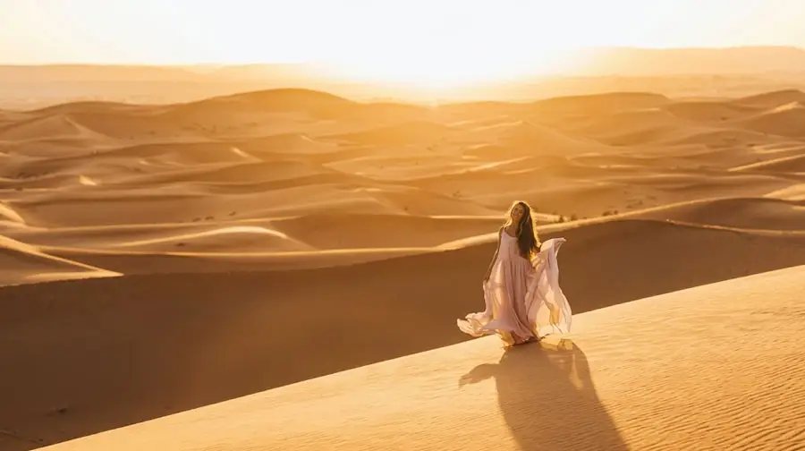 Woman in a flowing light-colored dress standing on a sand dune, enjoying the golden sunrise in the Abu Dhabi Desert.