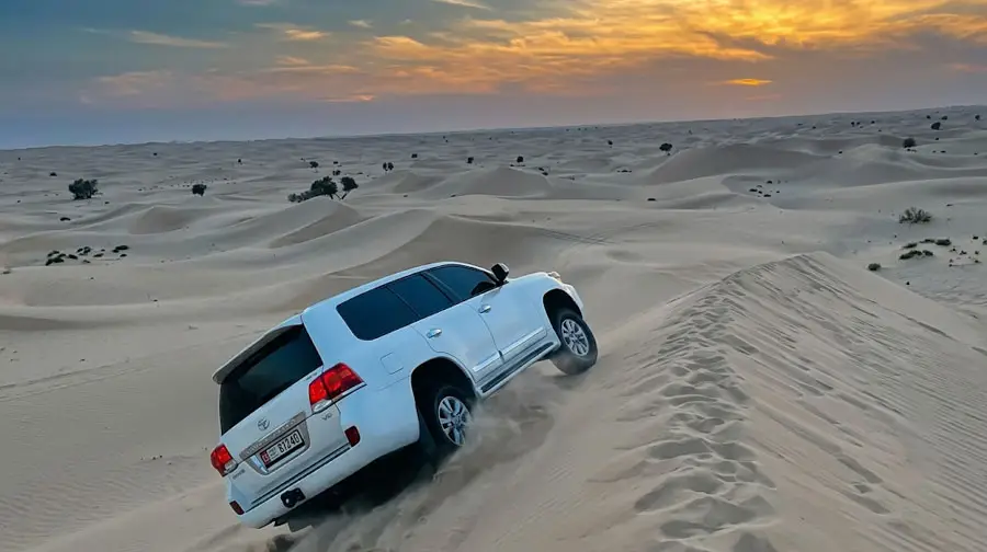 White Toyota Land Cruiser off-roading and climbing a steep sand dune, kicking up sand during a desert safari in Abu Dhabi at sunset.