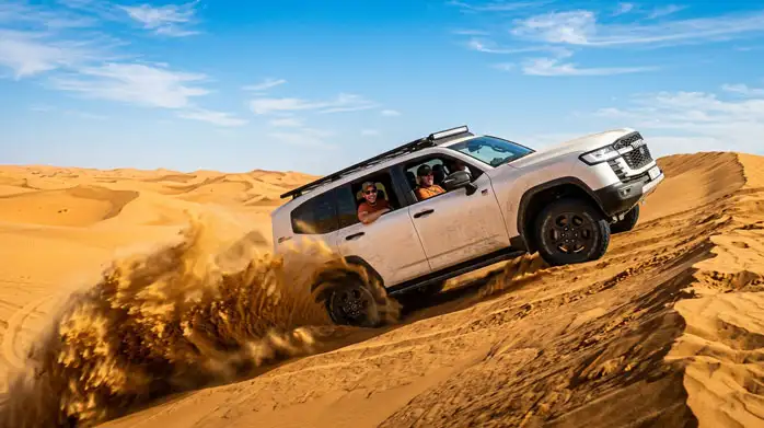A white 4x4 SUV kicks up sand while drifting across desert dunes under a bright blue sky.
